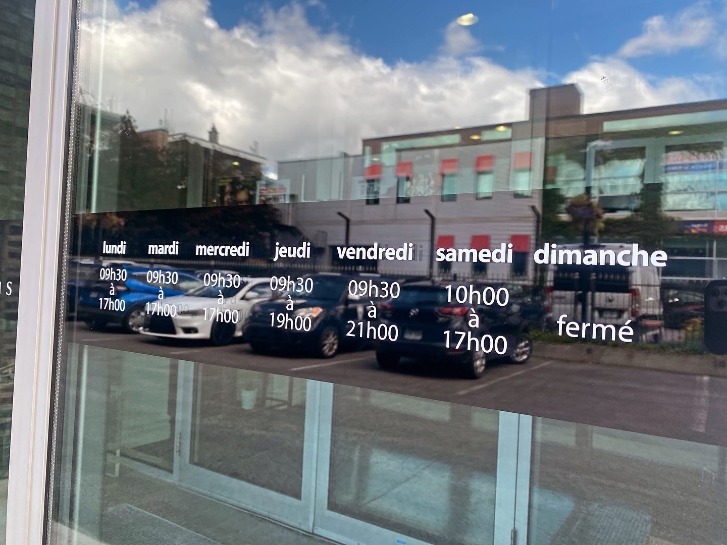 White opening-hours lettering on a window reflecting the street
